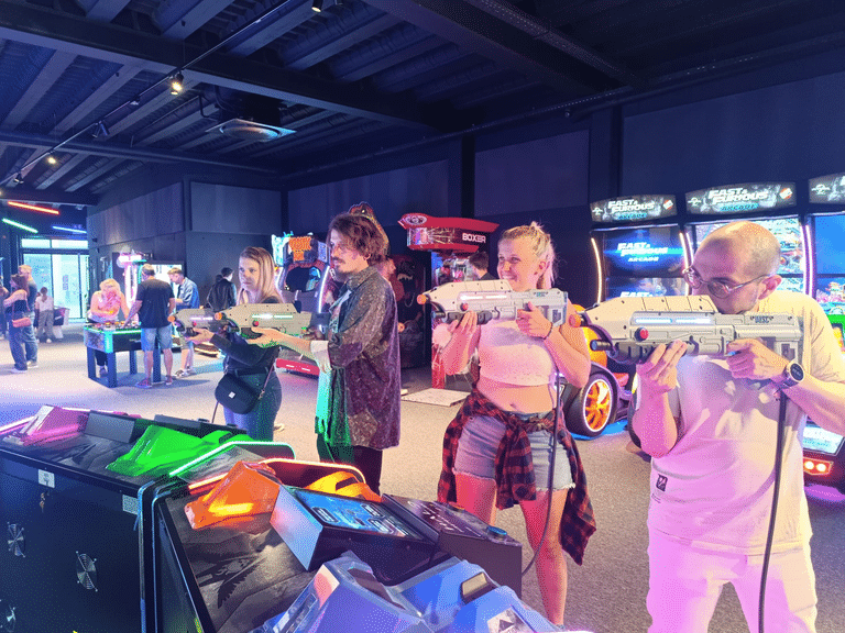 People in an arcade aiming large toy blasters at game cabinets in a dim, neon-lit space.