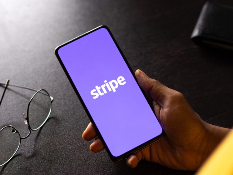 A hand holding a smartphone with an image of the "Stripe" logo in white on a purple background