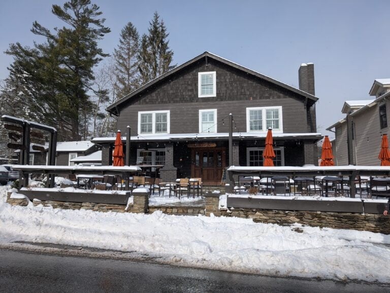 Hellbender Bed and Beverage, a cozy restaurant in Blowing Rock, NC, captured during winter with snow covering the ground. The building features a dark wooden exterior, outdoor seating with orange umbrellas, and a welcoming ambiance, showcasing one of the standout restaurants in Blowing Rock NC.