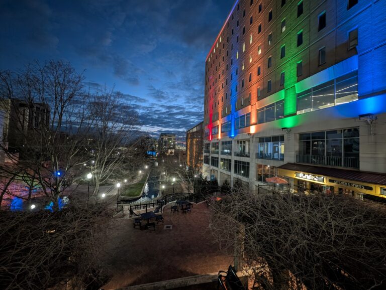 A vibrant evening view of Big Spring International Park in downtown Huntsville, Alabama. The sky is a deep blue with scattered clouds, while street lamps illuminate the park’s pathways. A modern building with colorful LED lighting in red, orange, green, and blue glows over the park, with a restaurant entrance visible on the ground floor. Featured image for the blog post '4 of the Best Hotels in Huntsville AL for Solo Travelers'.