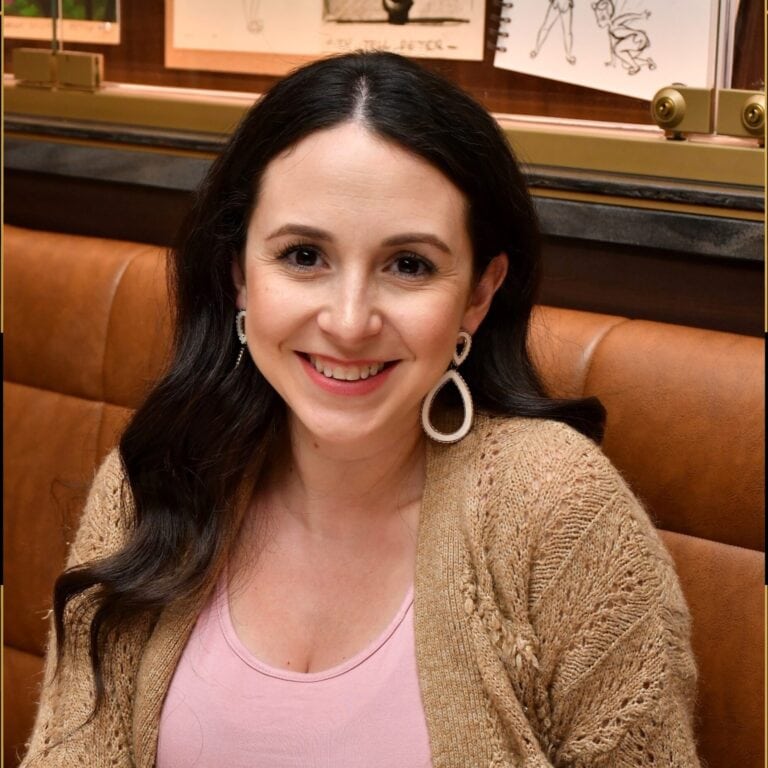 Smiling woman with dark hair wearing pink top and beige cardigan, enjoying a cozy restaurant setting.