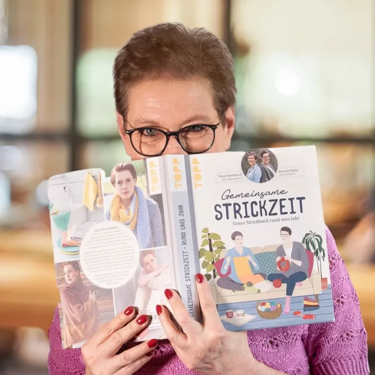 Frau schaut über ein Strickbuch in die Kamera