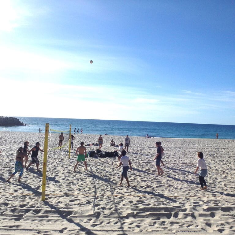 Menschen spielen Beachvolleyball an einem sonnigen Strand mit blauem Himmel und Ozean im Hintergrund.