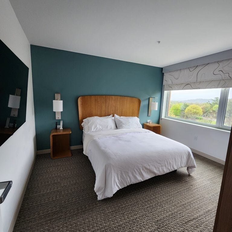A modern bedroom at The Element Hotel in Asheville, NC, featuring a comfortable queen-sized bed with white linens and a wooden headboard against a teal accent wall. The room includes matching bedside tables with lamps and a large window offering natural light and views of greenery outside. The clean, minimalist decor creates a serene and inviting atmosphere. Featured image for the blog post 10 Types of Solo Travel Accommodations (And How to Choose).