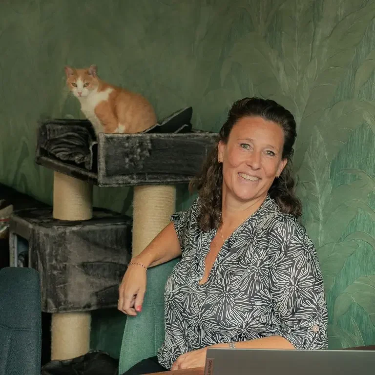 Mascha, de eigenaresse van Purrrfect by Mascha en kattenoppas.nl, zit glimlachend in een kamer met een kat op een krabpaal op de achtergrond.