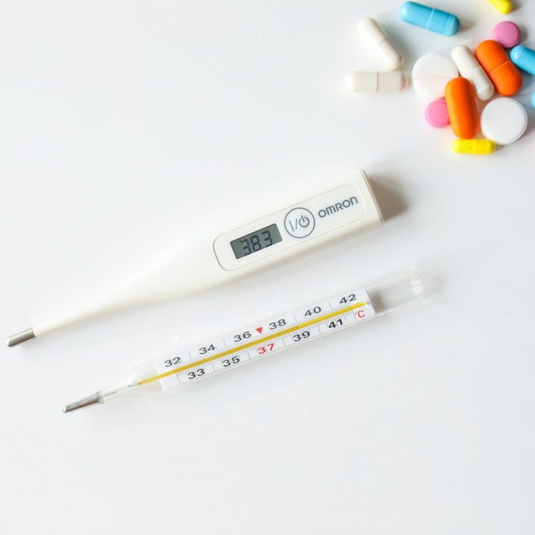 Ein digitales und ein analoges Fieberthermometer neben bunten Tabletten auf weißem Hintergrund.