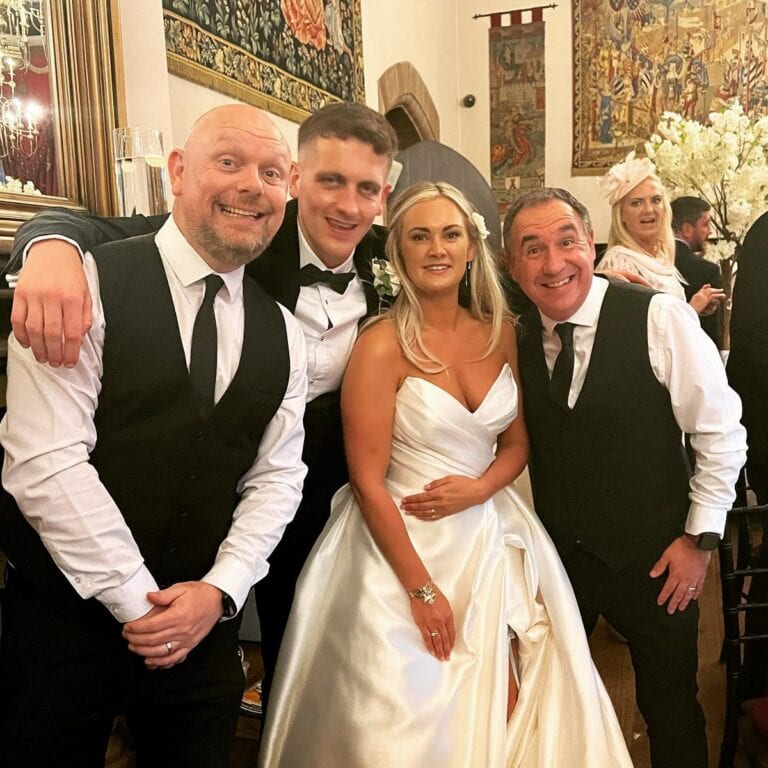The Sing Along Waiters performing at luxury castle wedding venue, award-winning singing waiters in formal attire with bride in elegant silk wedding dress, historic tapestry backdrop at prestigious UK venue, professional entertainment duo with 30 years experience