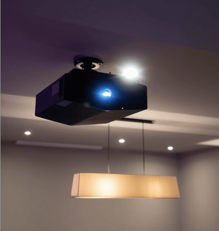 A ceiling-mounted projector is switched on, emitting a bright blue light in a dimly lit custom-built cinema room with pool table and bar in Fetcham, Surrey, featuring recessed ceiling lights and a rectangular pendant light hanging below.