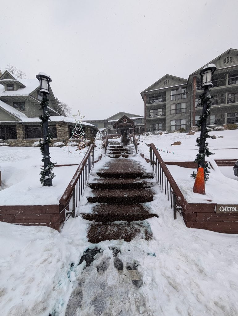 Snowy walkway leading to the main entrance of the Chetola Resort in Blowing Rock, NC, flanked by decorated lampposts, with the resort buildings in the background and a lighted Christmas tree display in the snow.