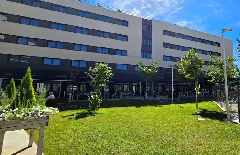 Edificio moderno con varias plantas y ventanas, rodeado de un jardín con césped, árboles y algunas personas sentadas. Torrejó