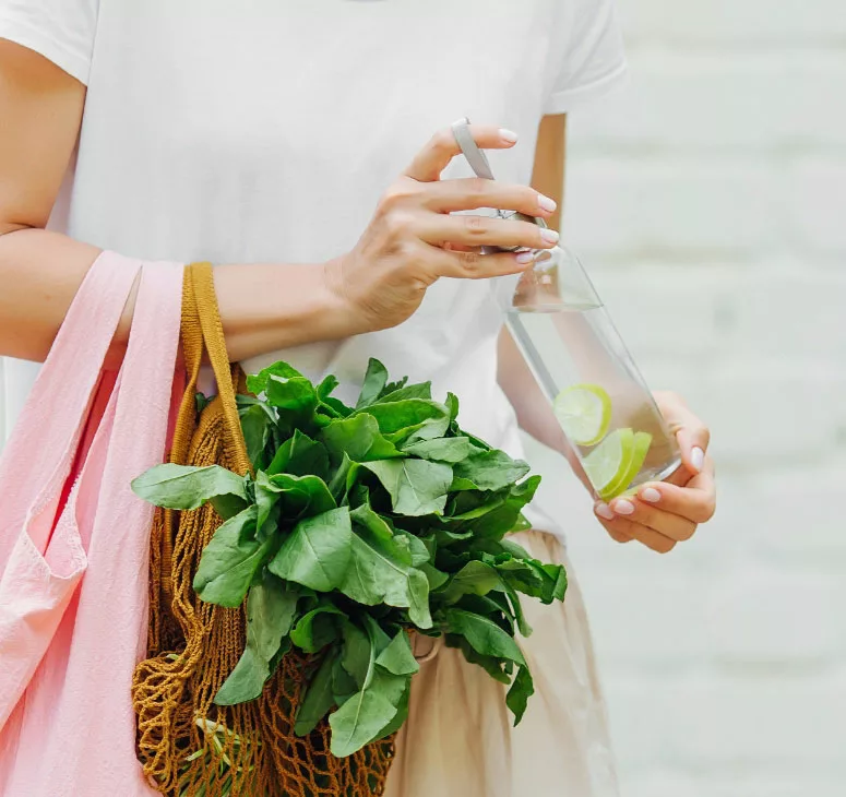Going-Greener-1 A woman holding a pink bag and a water bottle filled with water and sliced lemon.
