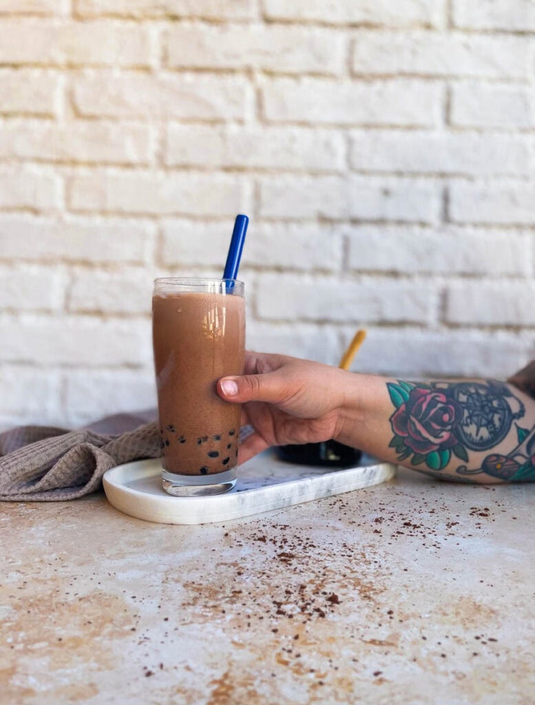 chocolate milk tea and hand reaching for it, boba pearls in a glass container behind, towel 