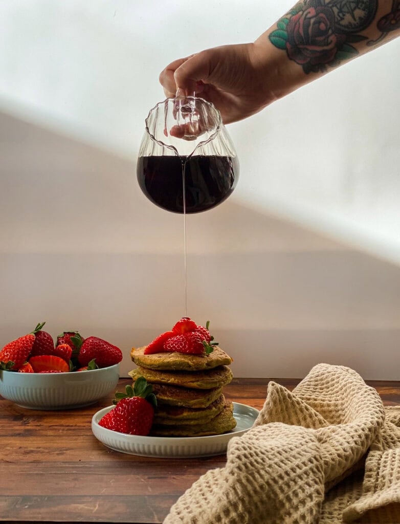 stack of vegan oat flour pancakes with maple syrup dripping on it from a jar held by a hand and strawberries