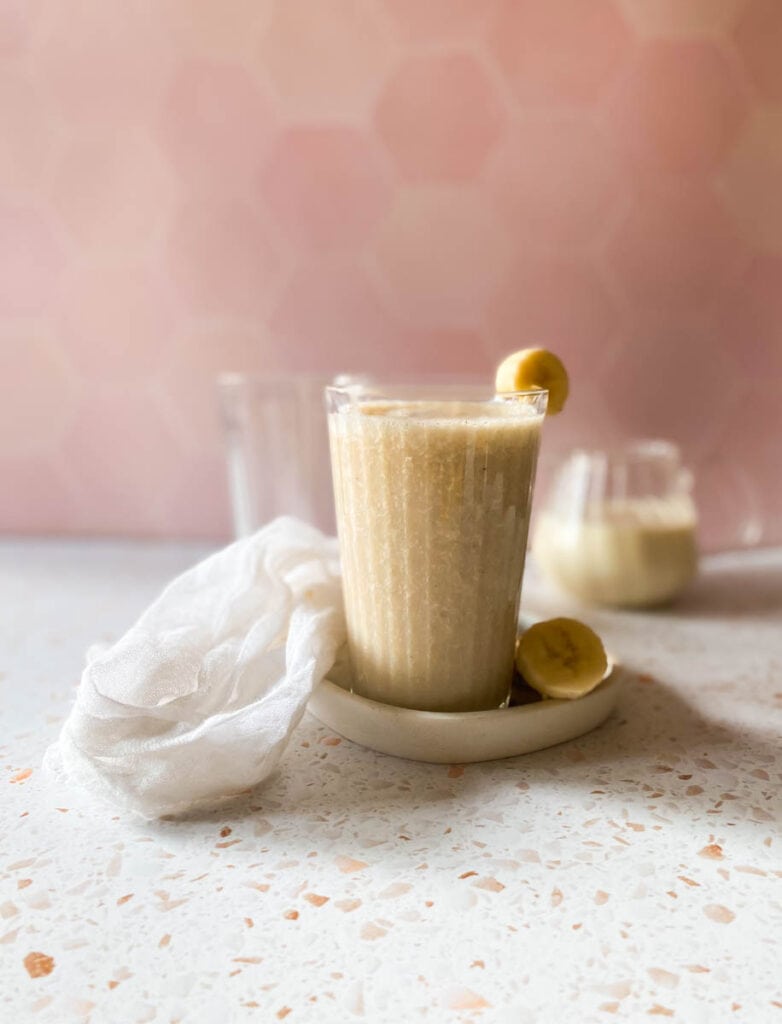 banana milk in a glass, on a tray with sliced banana, a towel, another glass and jug behind, a pink and speckled background