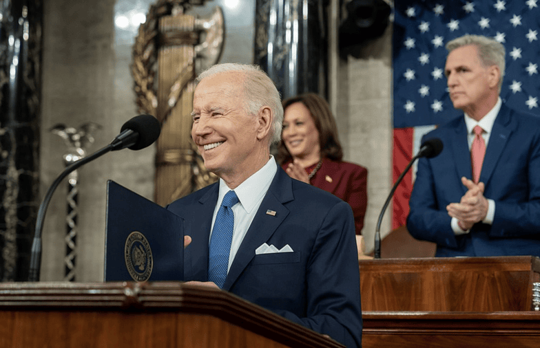 President Biden smiles from a lectern during the State of the Union as Vice President Harris and Speaker of the House McCarthy clap and look on.