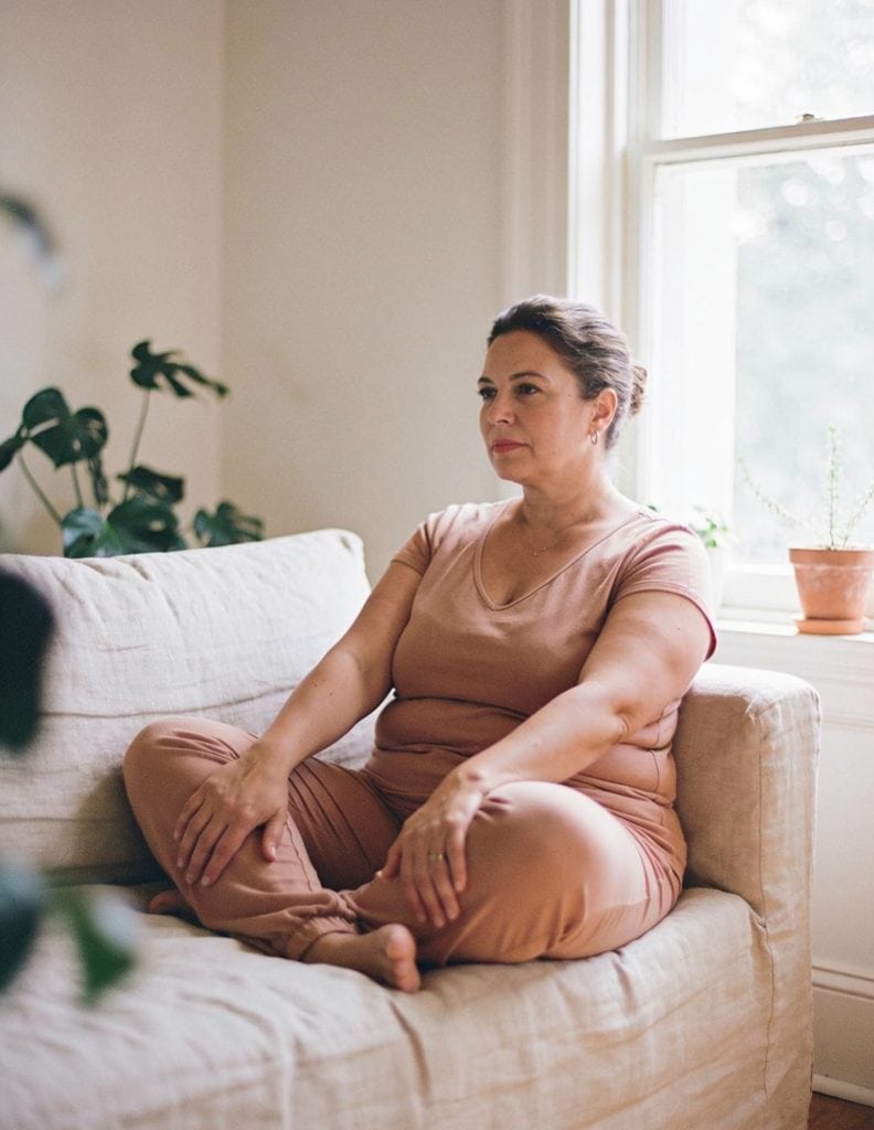 Mujer sentada en el sofá junto a la ventana, con las piernas recogidas y la mirada perdida