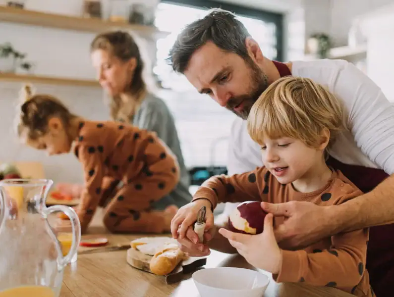 Pai ajuda menino a cortar uma maçã; ao fundo, a mãe ajuda outra criança a preparar uma receita.
