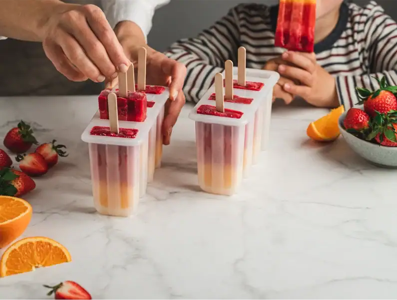 Adulto e criança a preparar picolés de fruta em moldes, numa atividade saudável na cozinha.