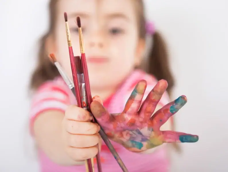 criança a segurar em pincéis e com as mãos pintadas.