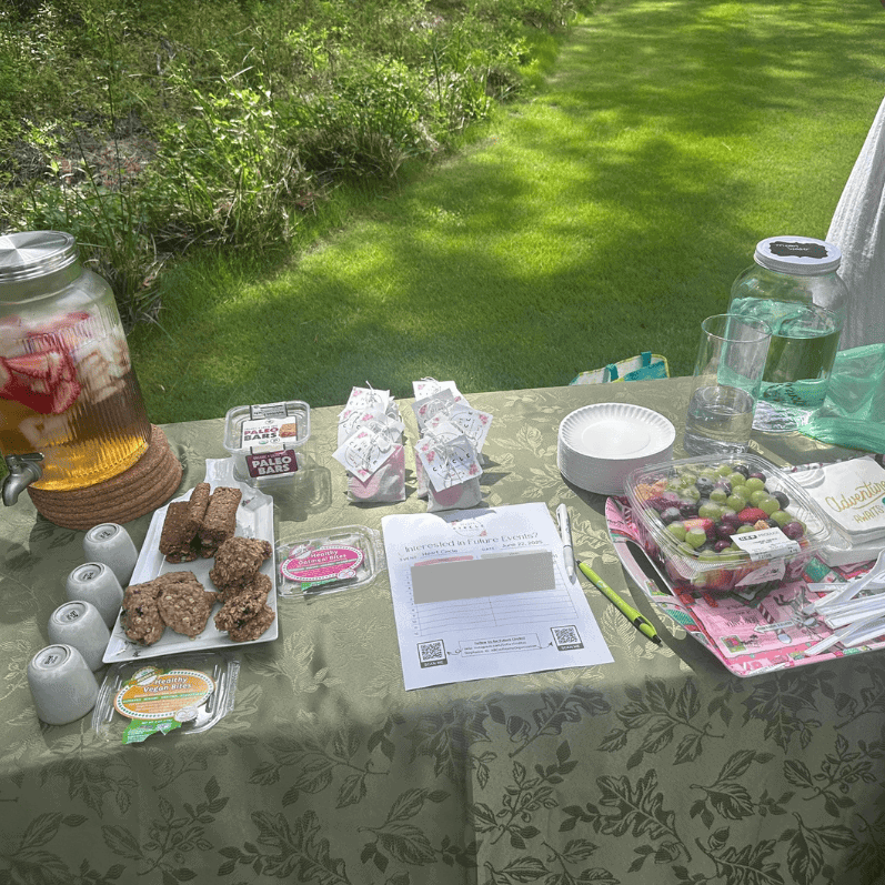 Vegan snack and beverage table set outdoors on a lush green lawn for a Soulstice Living event, featuring healthy treats, fruit, and refreshing drinks.