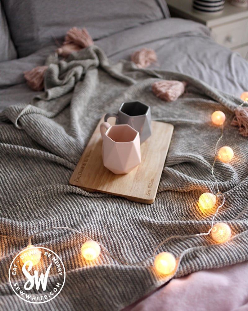 Cups of tea on a tray on a grey blanket on a bed with fairy lights. 