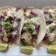 Fish tacos with flaky white fish, shredded cabbage, tomato, sauce, and shredded cheese sit on a plate with three lime wedges. Rubio's