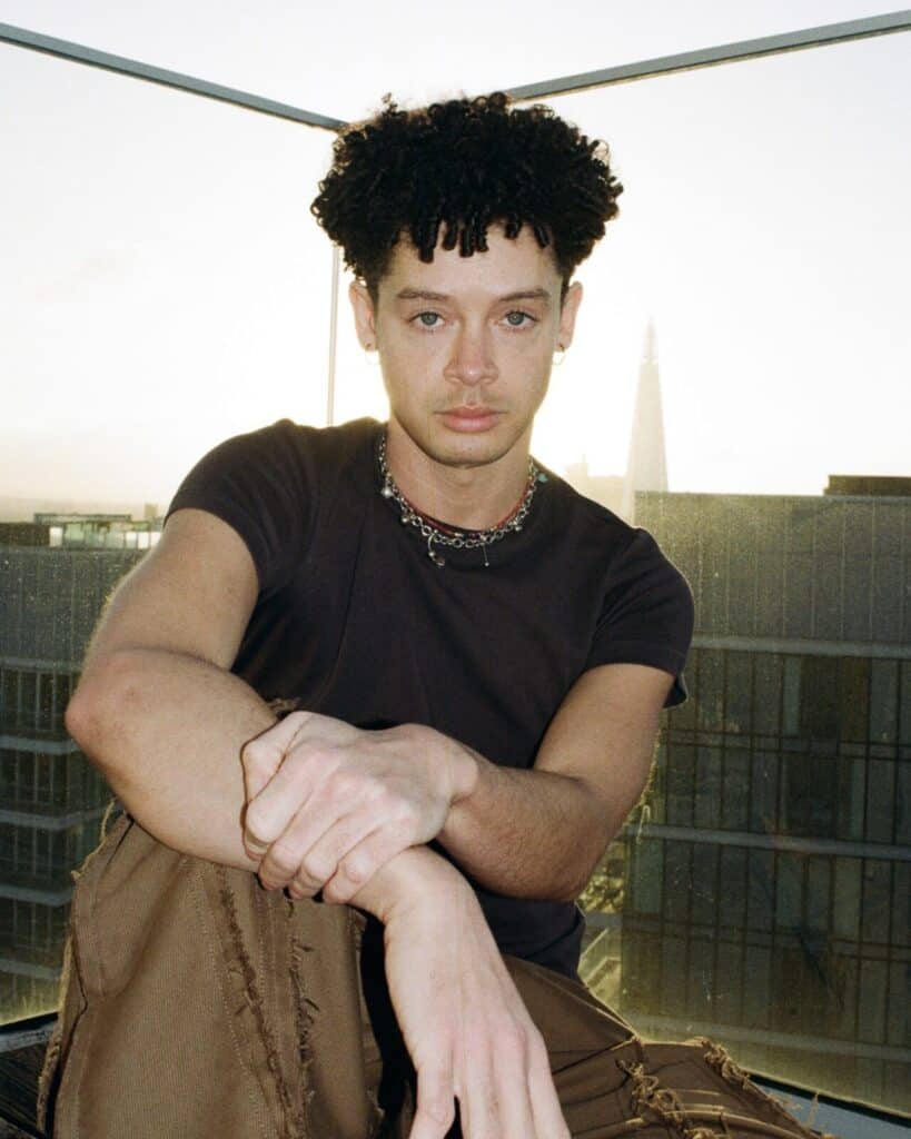 A person with curly hair sits on a rooftop at sunset, channeling Erykah Badu vibes in a black t-shirt, brown pants, and a chain necklace, with modern buildings and a glass railing in the background.