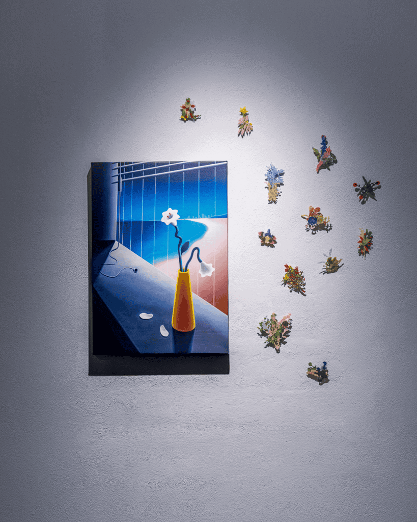 A painting by Hiroki Yagi of a yellow vase with white flowers sits on a dark table, hung on a light gray wall. Colorful, small, flower-shaped decorations are arranged on the wall to the right of the painting.