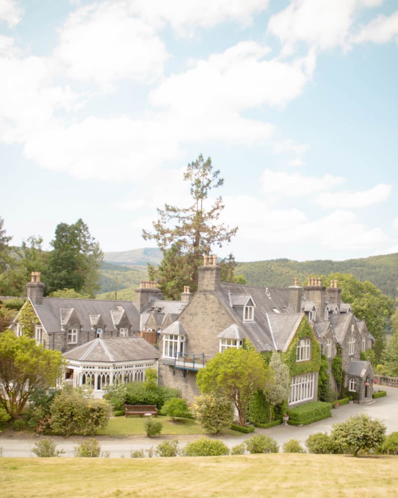 A grand stone manor house, Penmaenuchaf Hall Hotel, nestled among lush greenery and rolling hills in Wales. The historic building features ivy-covered walls, multiple chimneys, and a charming glass conservatory extending from one side. The surrounding gardens are beautifully landscaped, with manicured hedges, trees, and a wooden bench, creating a serene countryside retreat.