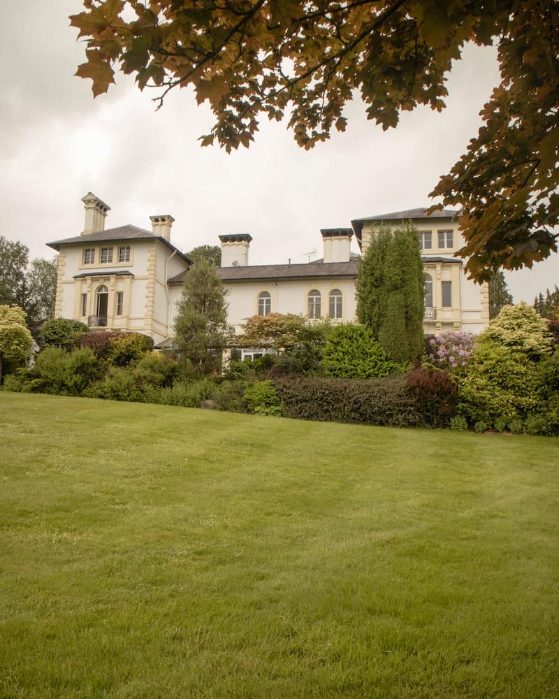 A grand country house hotel surrounded by lush green gardens and framed by autumn leaves. The elegant cream-colored building features tall chimneys, classic architecture, and well-manicured landscaping.