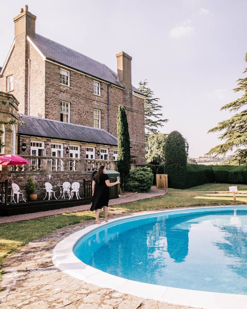 A woman in a black dress walks barefoot near a round outdoor pool, holding a glass of wine. The pool reflects the grand historic stone manor behind her, which features large windows, ivy-covered walls, and a charming terrace with white patio furniture. Lush greenery and rolling countryside complete the tranquil setting.