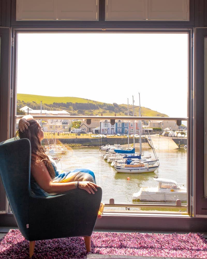 A woman in a blue and yellow dress relaxes in a cozy armchair by a large open window, gazing out at a picturesque harbor. Sailboats bob gently in the water, with colorful waterfront houses and rolling green hills in the background. Sunlight streams into the room, casting a warm glow on the serene scene.