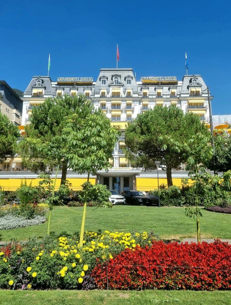 Elegantes Grandhotel Suisse mit gelben Markisen, blühendem Garten und blauem Himmel im Hintergrund.