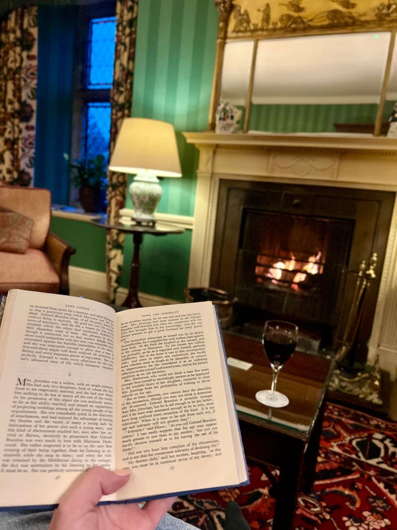 A person holds an open copy of Sense and Sensibility by Jane Austen, relaxing by a roaring fireplace. A glass of red wine rests on a wooden table, while the green-striped walls, vintage furnishings, and floral curtains create a warm, inviting atmosphere. The glow of the fire reflects in a large, ornate mirror above the mantel.