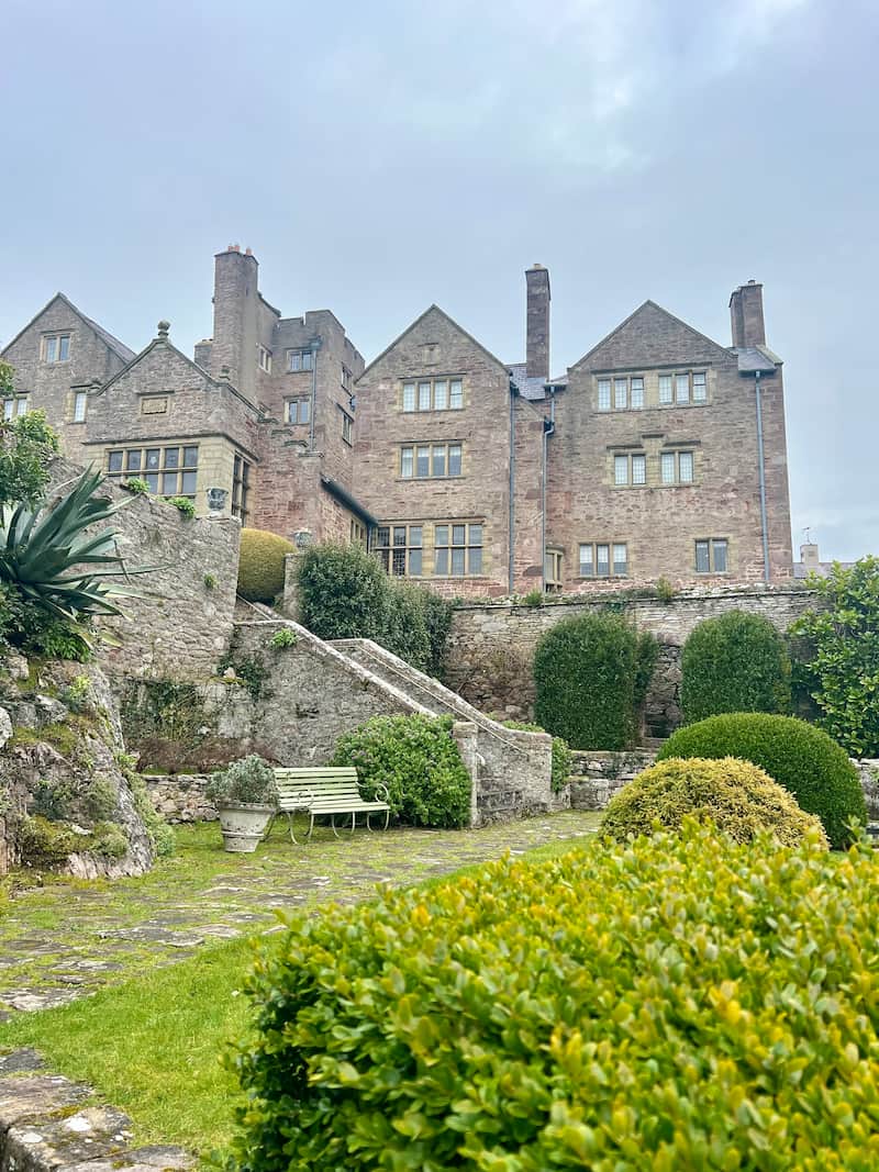 A historic stone manor house with tall chimneys and mullioned windows stands against a cloudy sky. The lush garden in the foreground features manicured shrubs, a stone staircase, and a weathered green bench nestled among ivy-covered walls. The scene has a timeless, tranquil feel.
