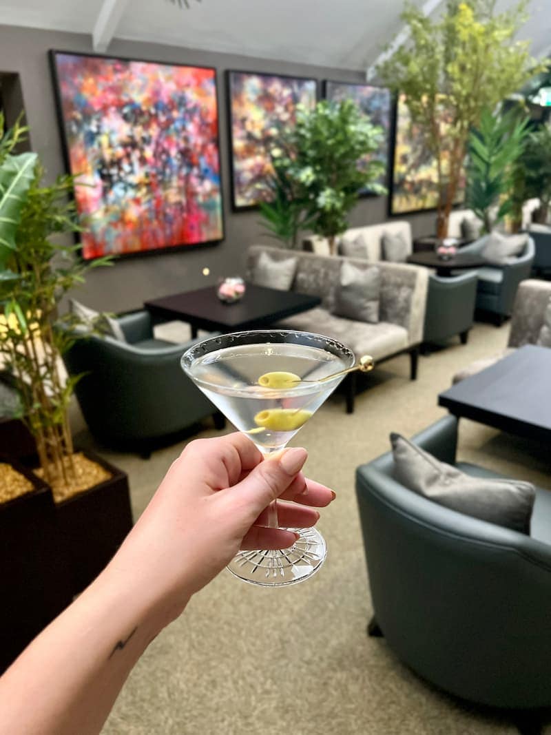 A hand holding a classic martini with two green olives on a cocktail pick, set against the stylish lounge of Twr y Felin Hotel. The modern space features plush seating, lush greenery, and vibrant abstract artwork on the walls. The setting exudes elegance and sophistication.