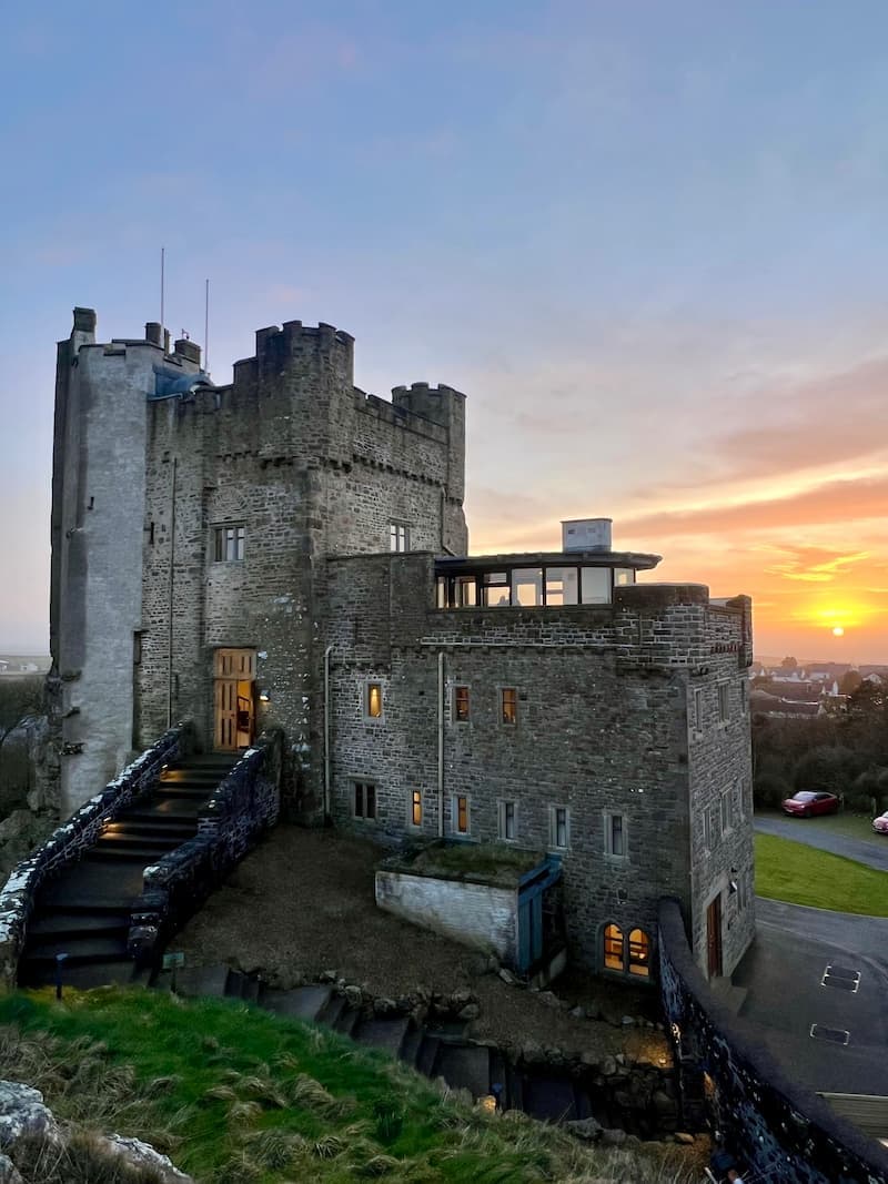 Roch Castle in Wales at sunset, with its medieval stone structure perched atop a rocky hill. A warmly lit staircase leads to the entrance, contrasting with the rugged exterior. The sky is painted in hues of orange and pink as the sun sets over the landscape, casting a golden glow on the castle’s modern glass extension.