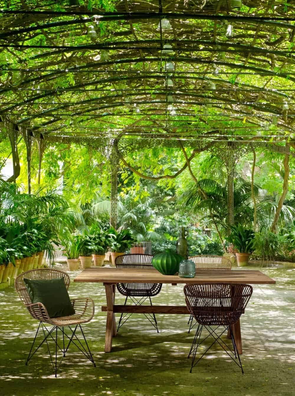 Ampia area patio con tavolo naturale, piante lussureggianti e copertura in foglie verdi per ambienti rilassanti.