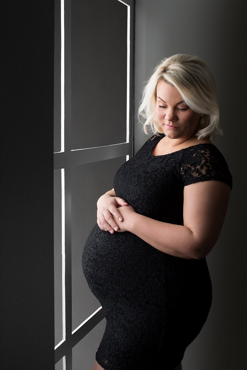 Elegante gravidfotografi av gravid kvinne i svart blondekjole, nyter stille øyeblikk foran vindu, perfekt for prenatal fotoshoot og minner til den kommende barnetiden.