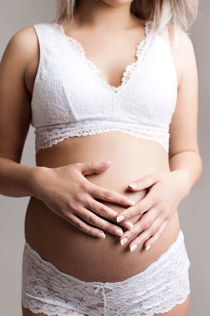 Naturlig gravidfoto av mamma med mage i fokus, intim gravidbilder i hvit blondetopp og undertøy, graviditetsshoot for forventningsfulle mødre, vakker gravidfotografering, gravidfotografering i Norge.