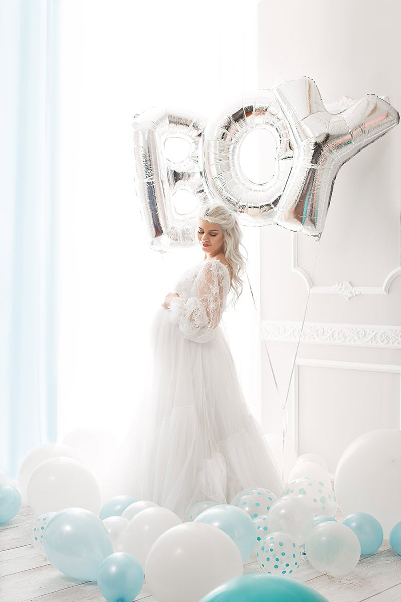 Bilda av en gravid kvinne i hvit kjole med ballonger og "BOY" luftballonger i bakgrunnen, perfekt for gravidfotografering.