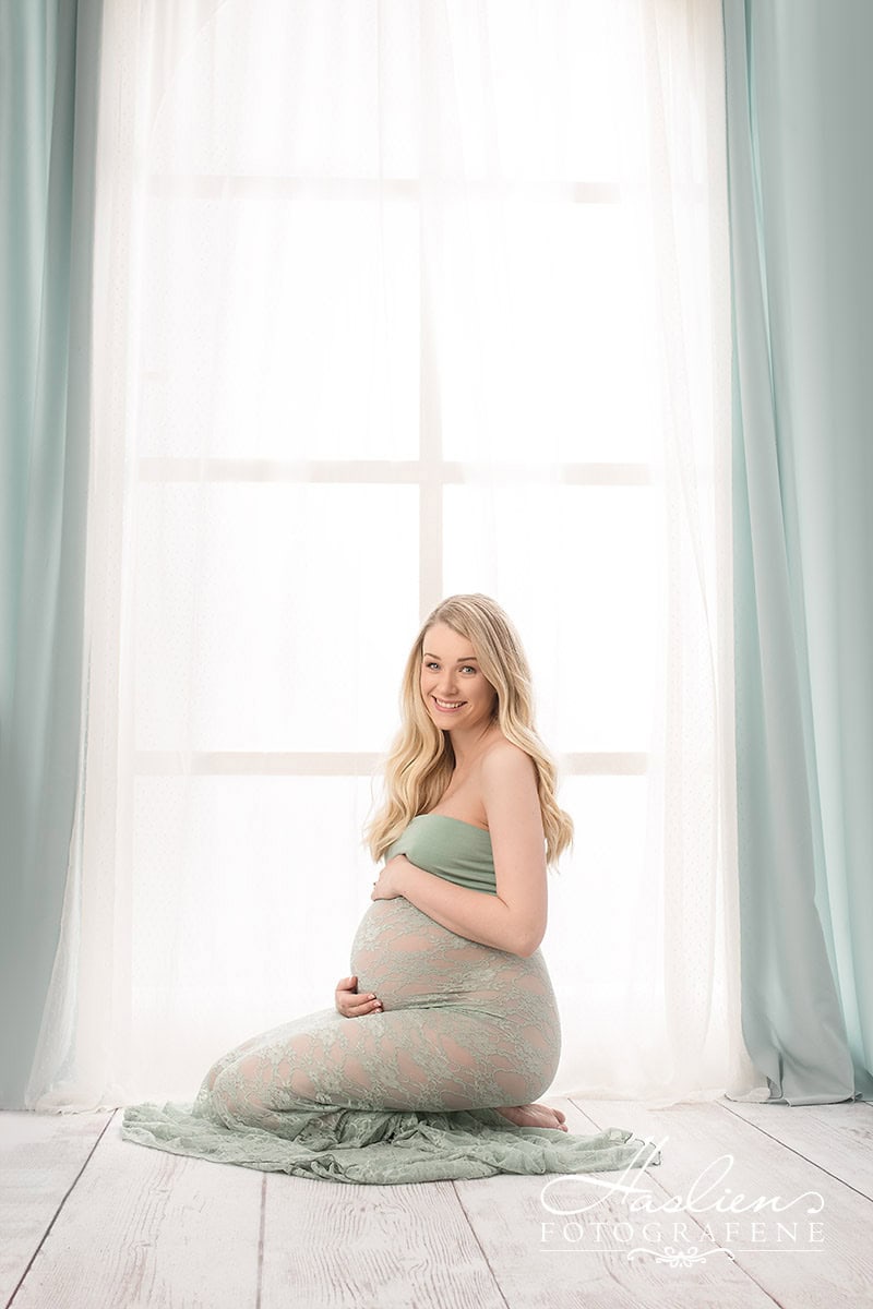 Kvinne i graviditetsshoot, sitende på huk foran et stort vindu med myke gardiner, smilende og glad, rolig og harmonisk atmosfære, vakkert lys, gravidfotografering, portrett av gravid kvinne, eleganse og graviditetspanorama.