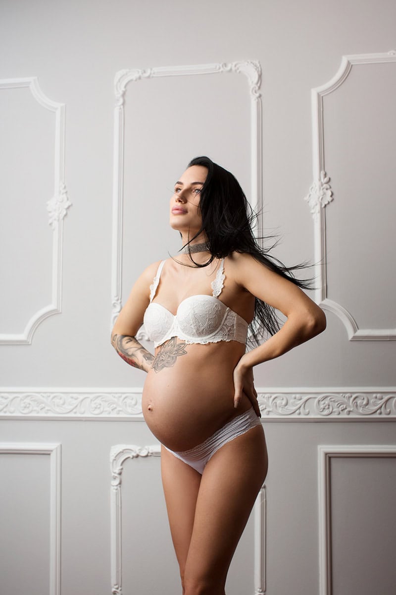 Unge gravid kvinne i hvit undertøyposerer i elegant rom med hvite veggdekorasjoner, fremhever den naturlige skjønnheten og styrken i graviditeten, perfekt for gravidfotoshoot.