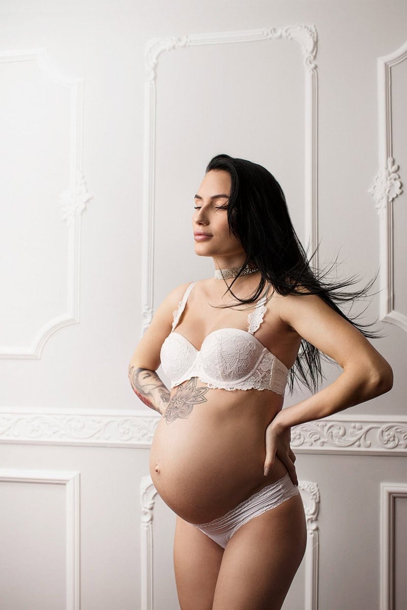 Kvinne med gravid mage i lingerie, fotografert i hvitt rom med elegant dekor, for gravidfotografering og gravidfoto.