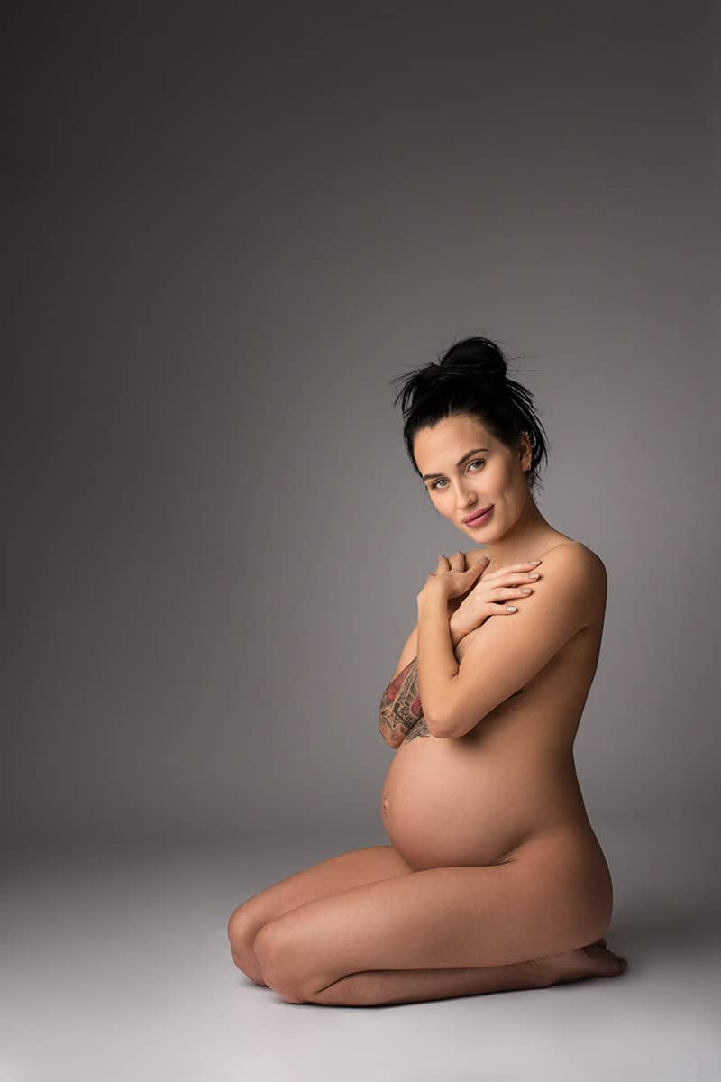 Naturlig gravidfoto av kvinne med tatovering, som smiler rolig mot kamera, fremhever graviditet og kvinnelig skjønnhet, fotografert i studio med enkel grå bakgrunn.