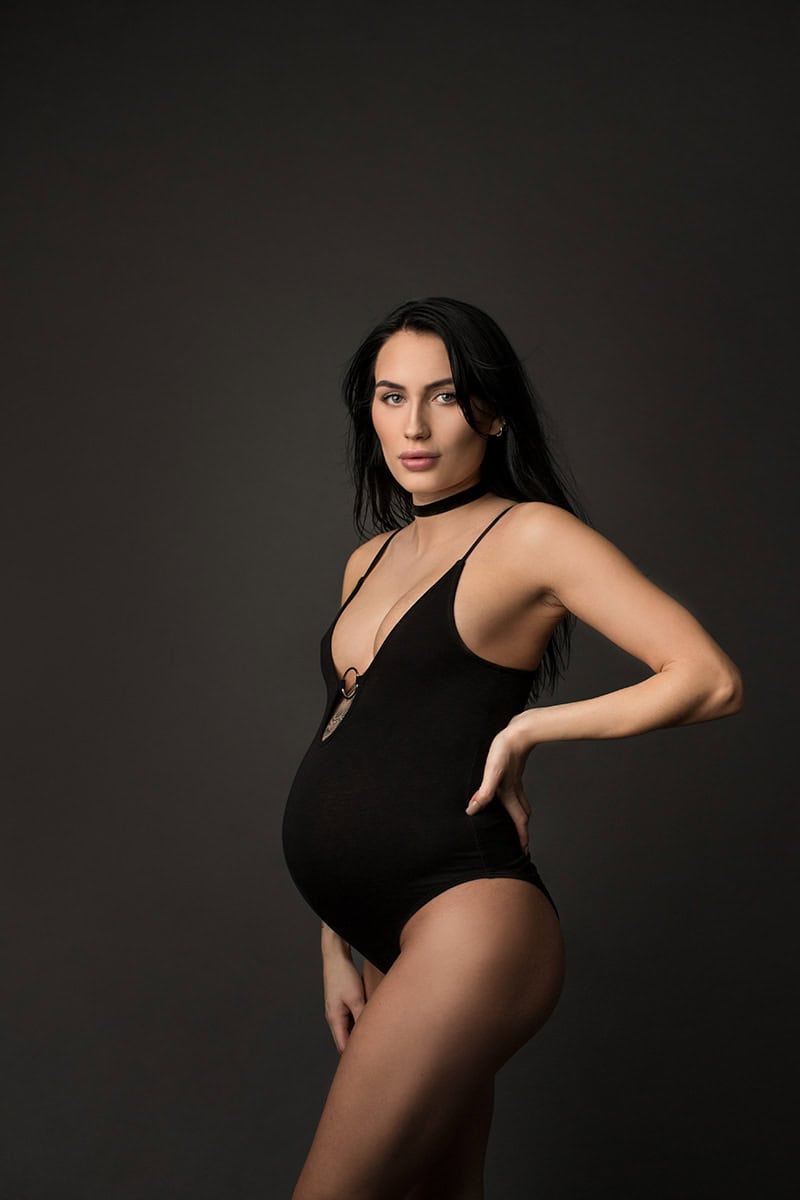Elegant gravidfoto av en gravid kvinne i svart undertøy, fotografert mot en nøytral mørk bakgrunn, fremhever graviditet og kvinnelig skjønnhet. Fotografering for gravidbilder, gravide fotoshoots, og morskapstraumer.