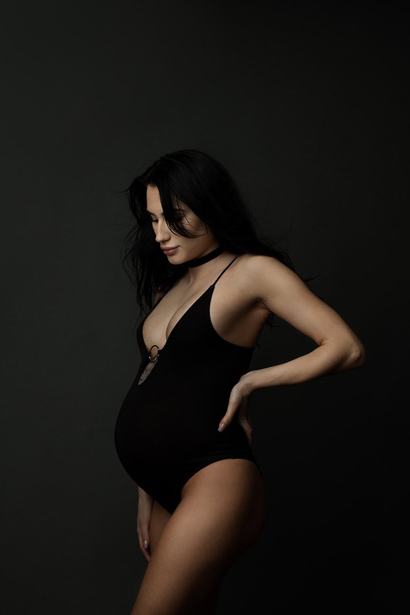 Kvinne med gravidmage i svart undertøy, fotografert i studio mot svart bakgrunn for gravidfotografering, vekt på graviditet og vakker kvinnelig form.