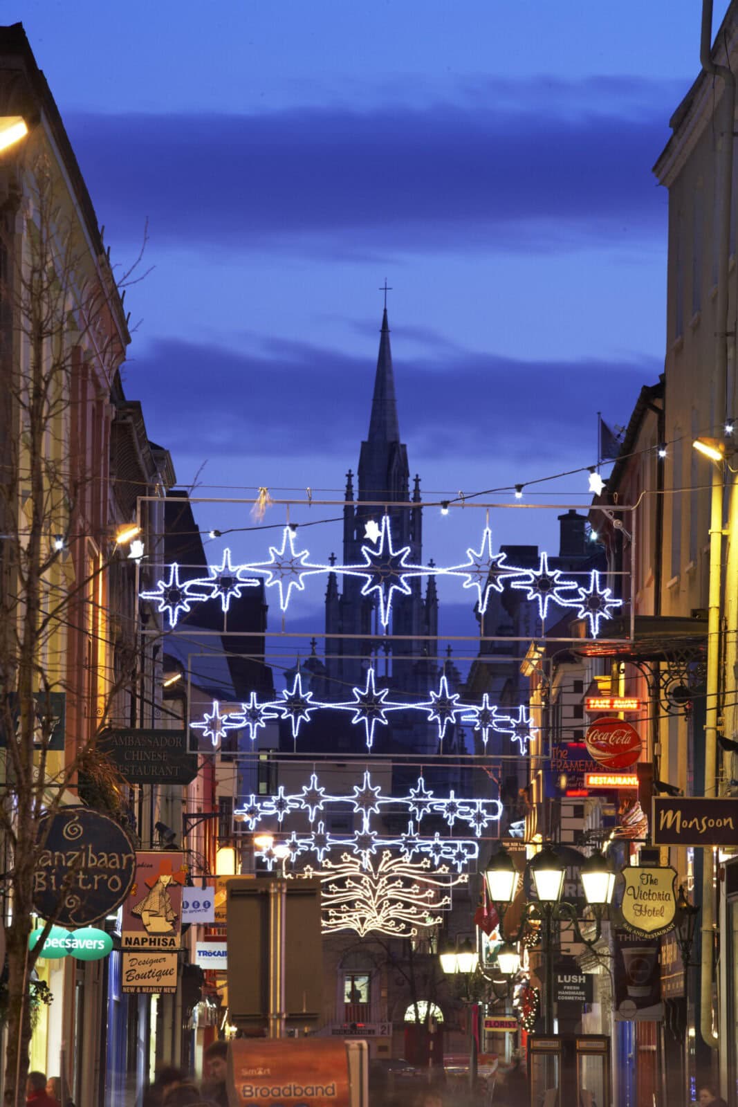 Beleuchtete Einkaufsstraße mit festlicher Weihnachtsdekoration und einer Kirche im Hintergrund bei Dämmerung.