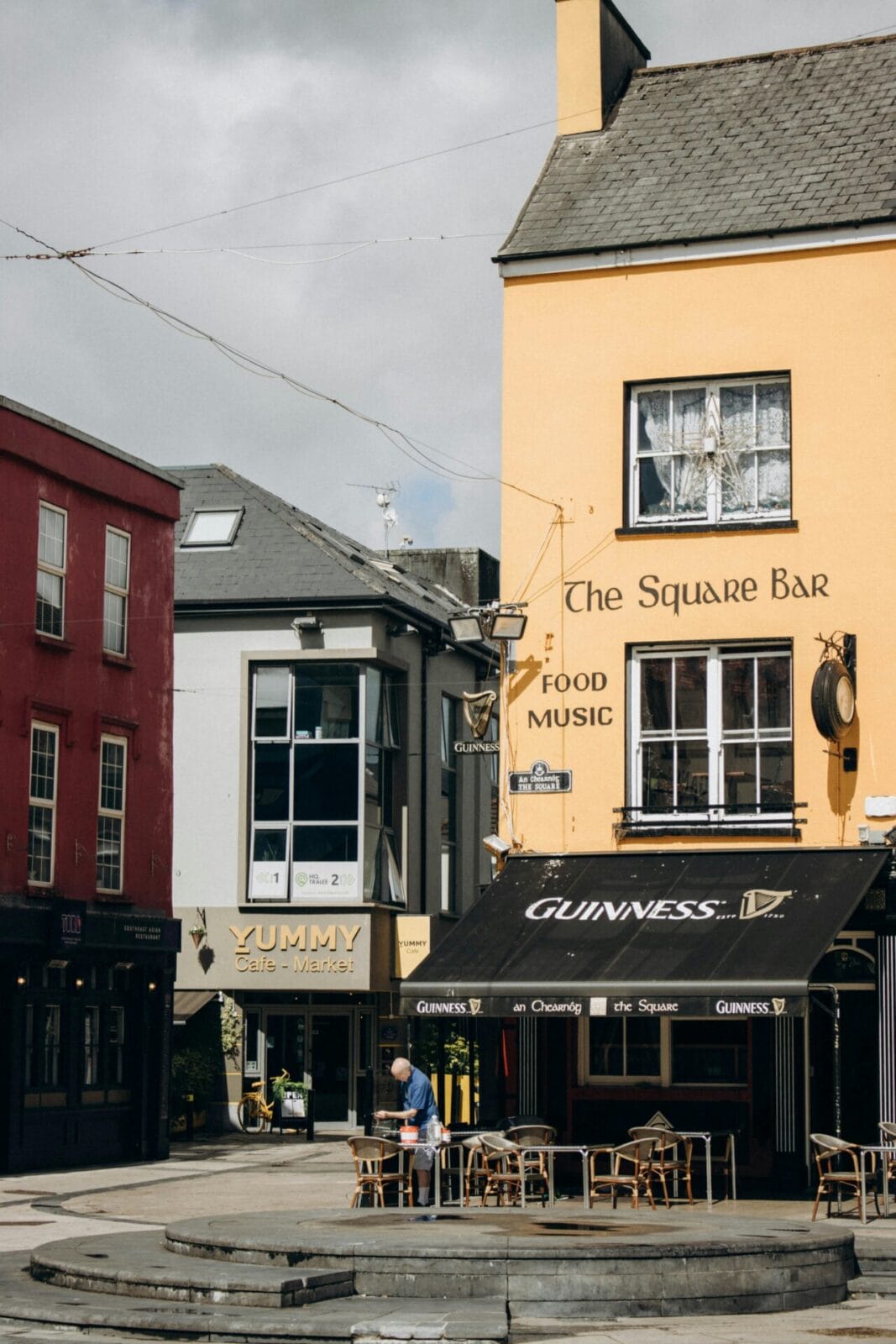 Charmanter Platz mit bunten Gebäuden, Café und traditioneller Bar mit Guinness-Markise in einer irischen Stadt.