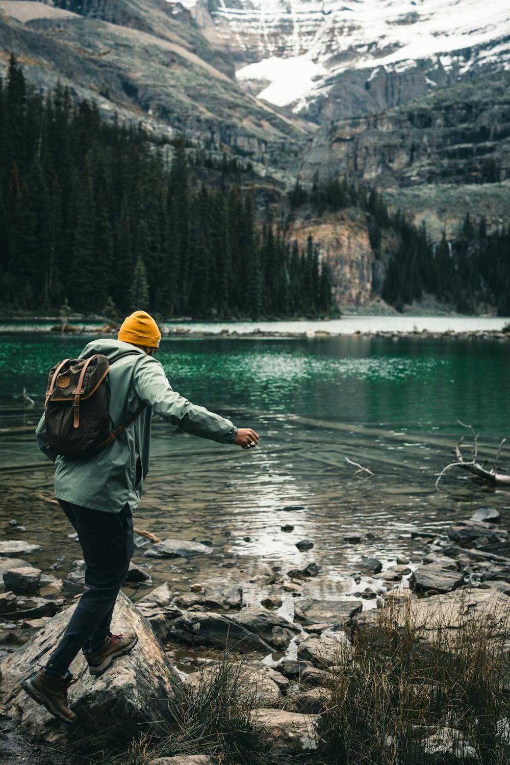 Ein Wanderer mit Rucksack und gelber Mütze erkundet einen Bergsee mit klarem Wasser, umgeben von Tannen und schneebedeckten Gipfeln.